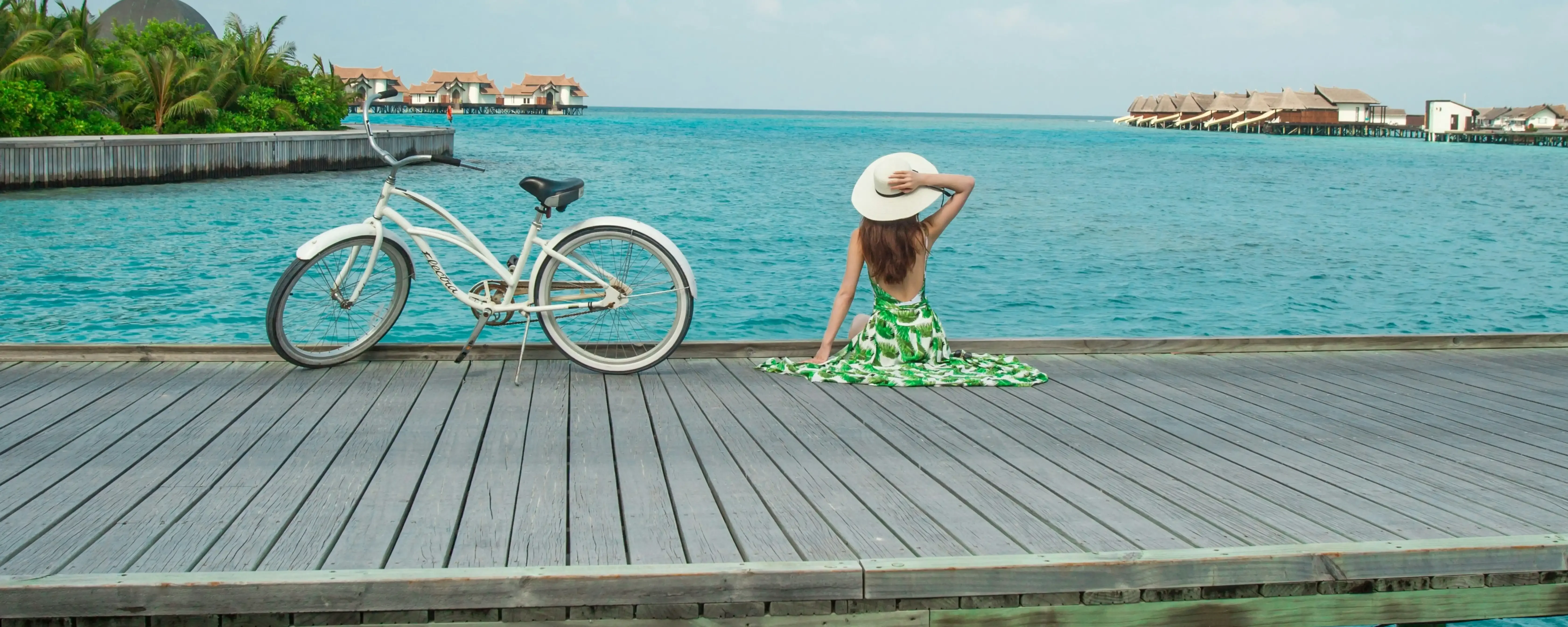 Girl sits on boardwalk looking at water and bungalows, bike near, Maldives tour.