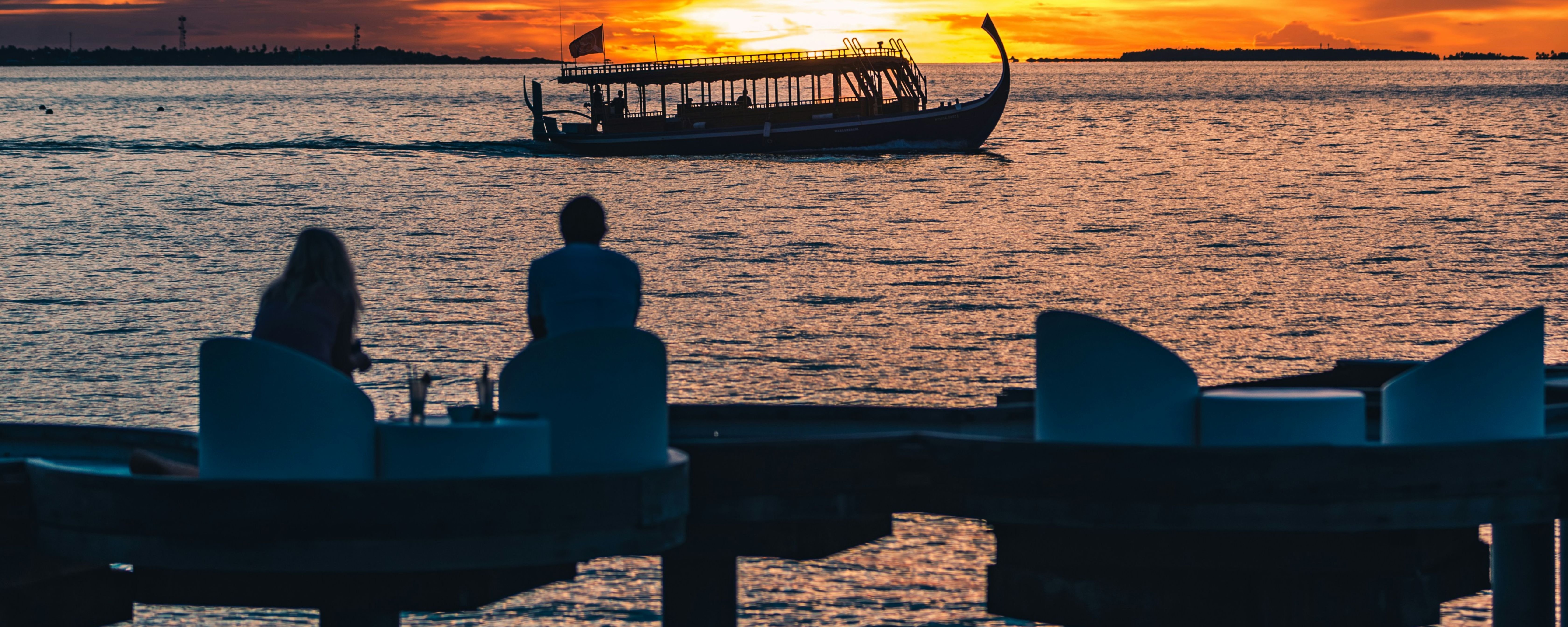 Two people watch the sunset over the sea, a boat in the distance, on a Maldives tour.
