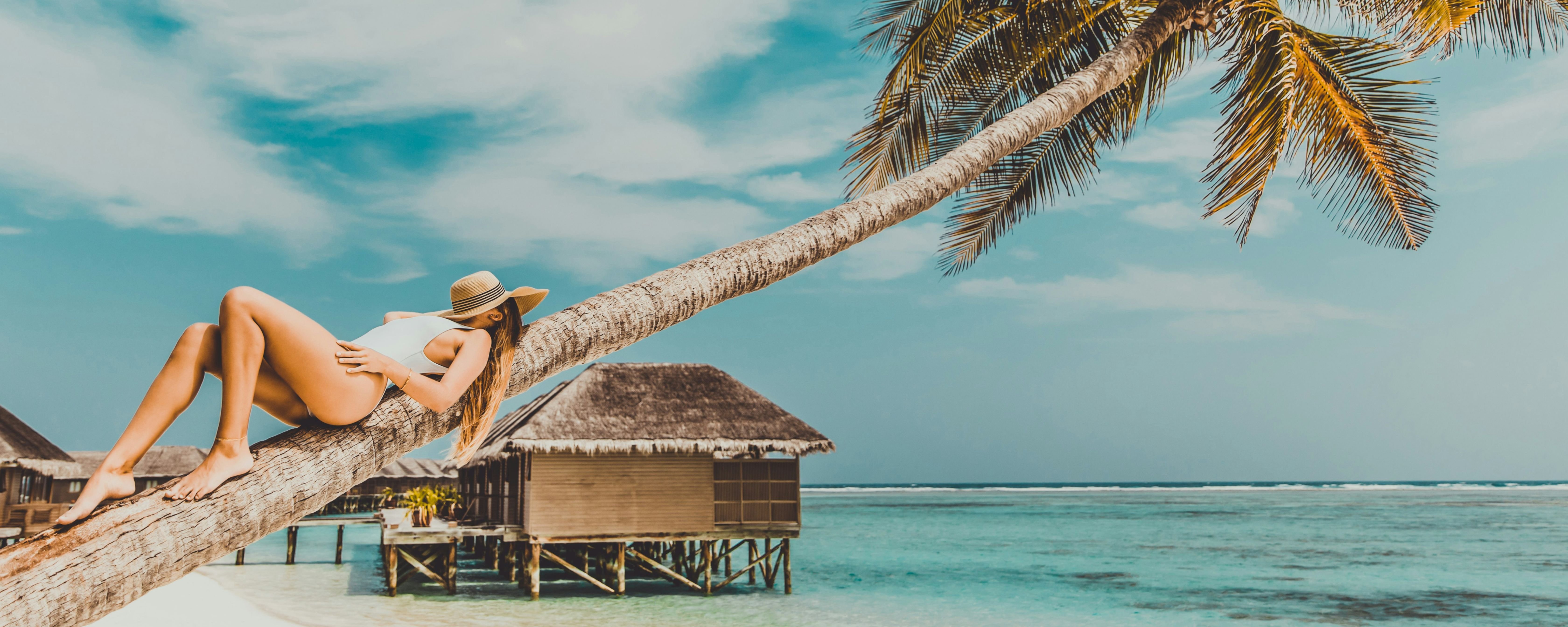 Maldives tour package: Woman in a white swimsuit and hat sitting on a leaning palm tree by the turquoise water.