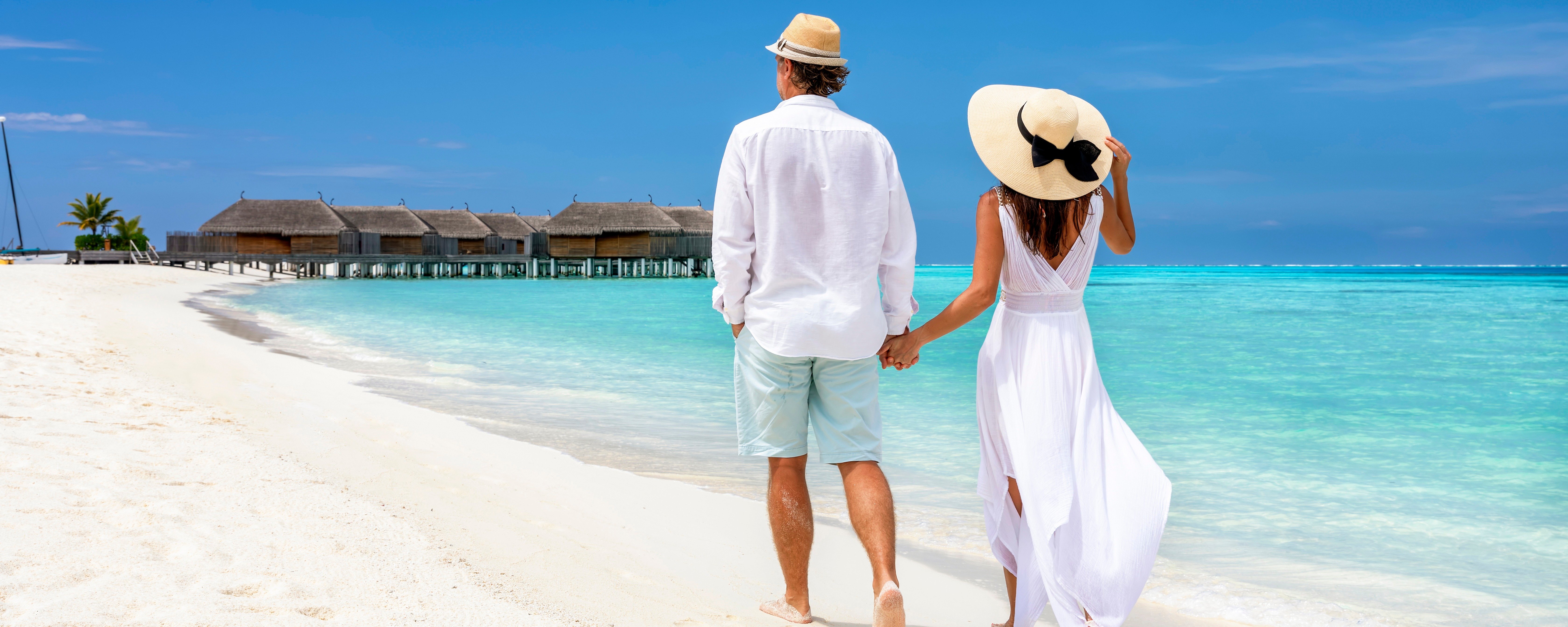 The image shows a couple on a white-sand beach with clear turquoise water and overwater bungalows in Maldives tour.