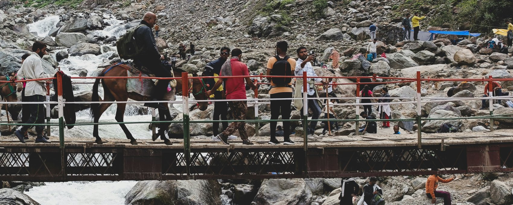 Pilgrims on Kedarnath trek, doing Char Dham Yatra. Mules help cross river bridge. Scenic view.
